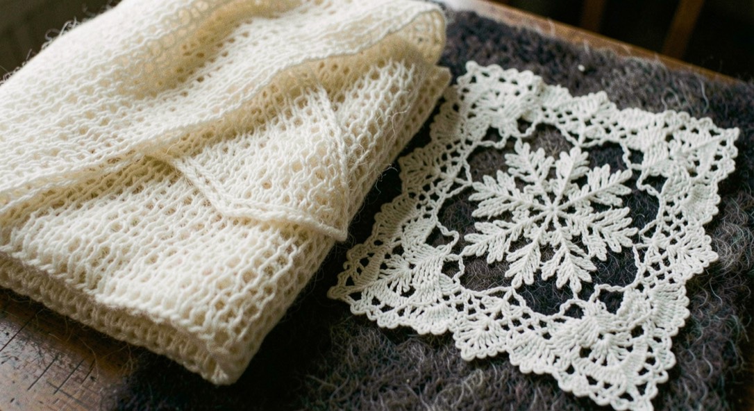 Cream gossamer knitted down shawl folded light as air beside a square of dense white bobbin lace with snowflake pattern on dark wool